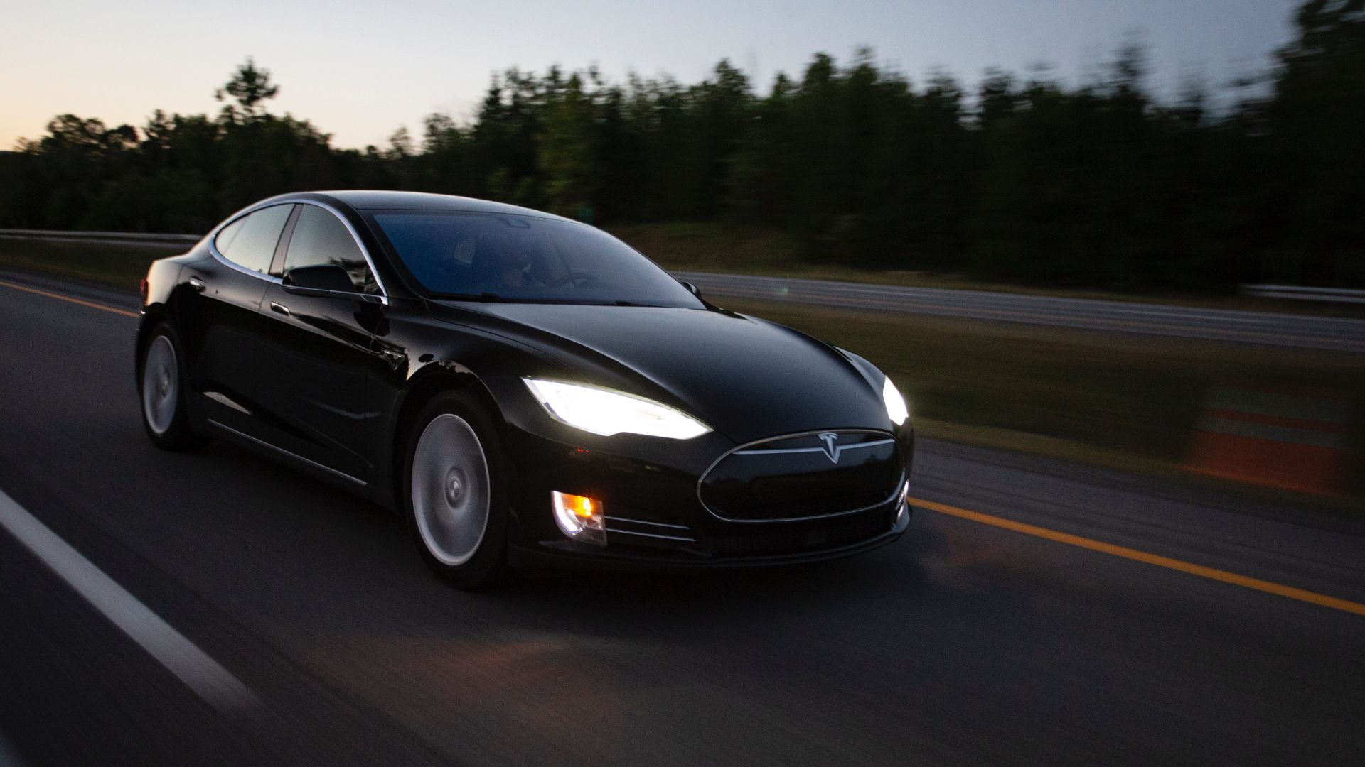 a black car driving down a road next to a forest.|a white electric car driving down a highway.