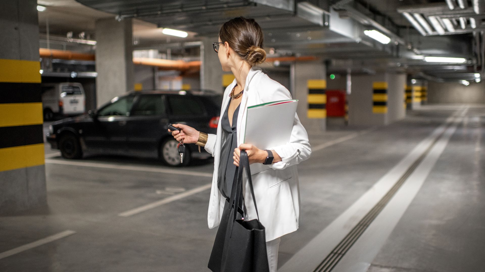a woman standing in a parking garage holding a clipboard.|a man sitting in a car talking on a cell phone.|a woman driving a car wearing glasses and a sweater.