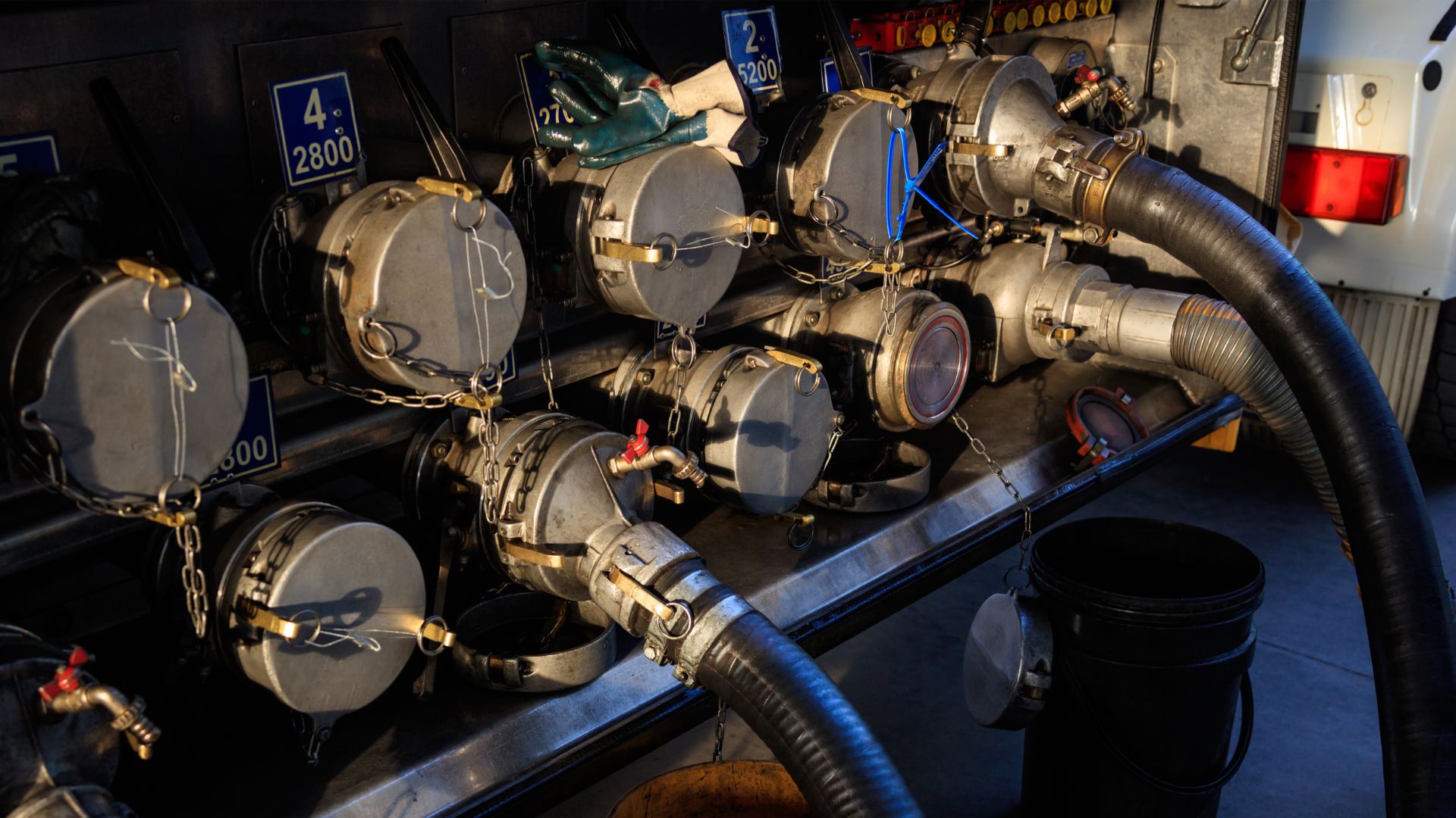 a bunch of pipes are connected to the back of a truck.|a group of plastic containers sitting next to each other.|a forklift with blue barrels on a pallet.|a close up of a person holding a gas pump.|a close up of a truck with hoses attached to it.