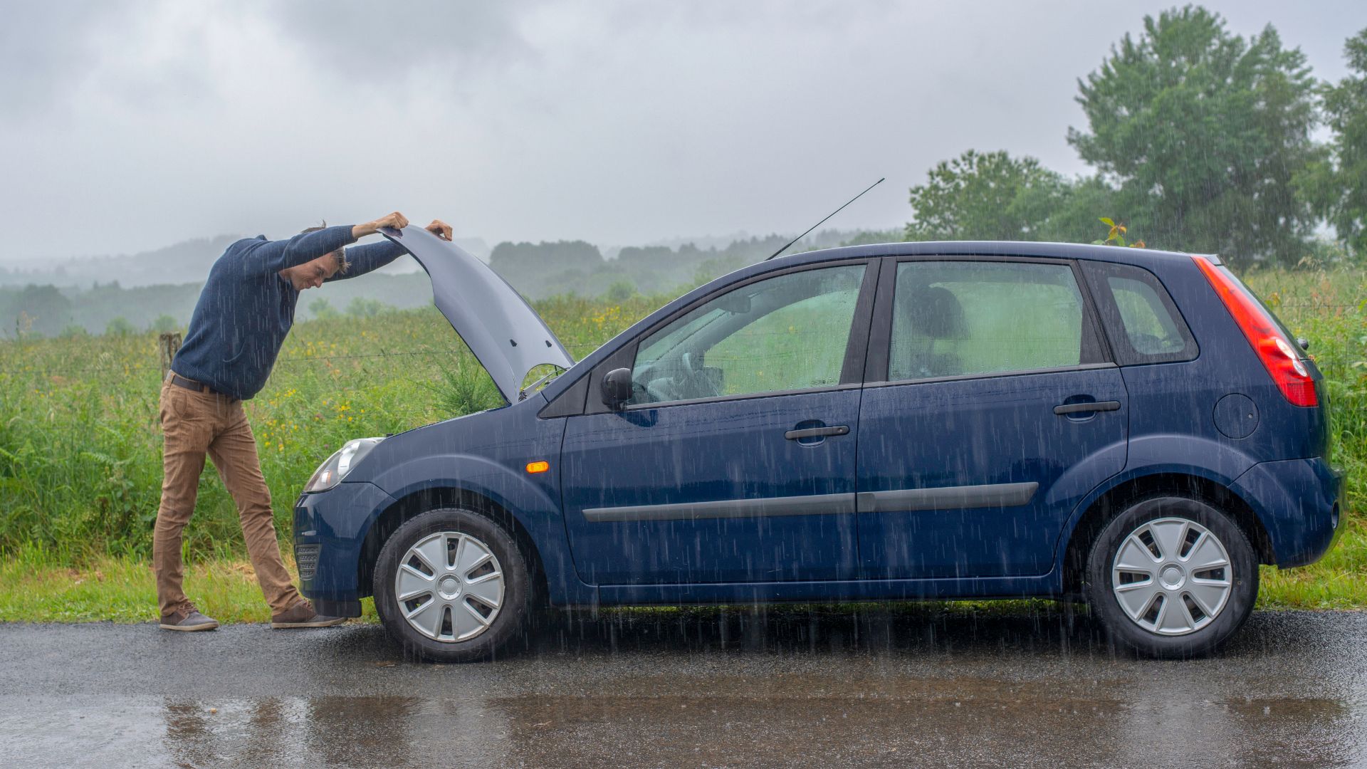 a man standing next to a blue car in the rain.