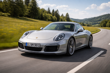 A silver Porsche 991 sports car drives on a winding country road, surrounded by green fields and trees under a partly cloudy sky.