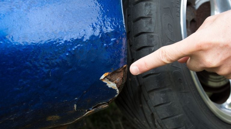 a person points at a piece of rust on the side of a blue car.