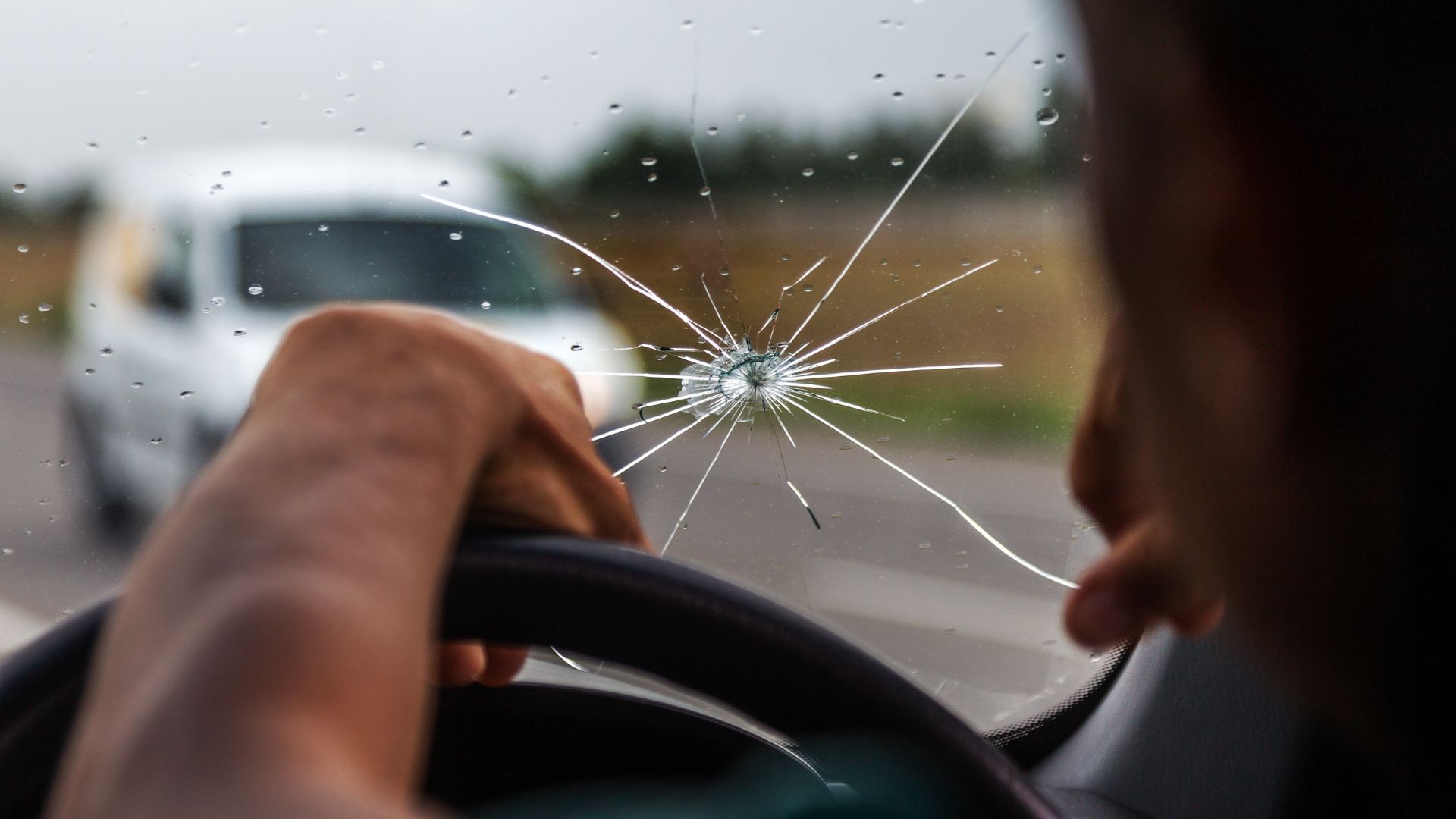 a person holding the steering wheel of a car with a broken windshield.